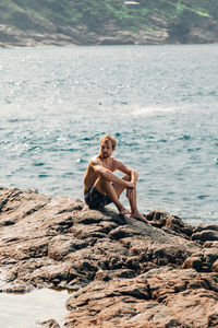 Full length of shirtless man sitting on rock at beach