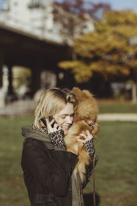 Smiling blond woman talking on mobile phone while carrying pomeranian at park on sunny day