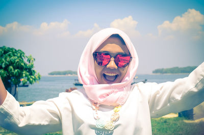 Front view of happy young woman wearing sunglasses at lakeshore against sky