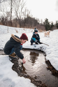 Two boys playing in a puddle on a snowy path with dog in background.