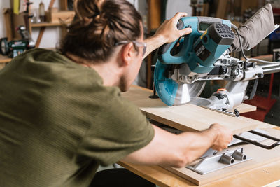 Back view of male carpenter in protective goggles cutting wooden plank with miter saw while working in bright workshop