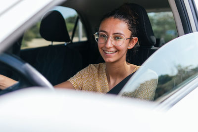 Young happy female in casual clothes and glasses looking at camera and driving contemporary vehicle on sunny day