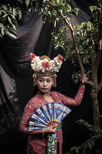 Woman in traditional clothing holding hand fan while standing by plants