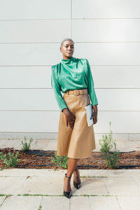 Full length of serious young african american businesswoman with short dyed hair in fashionable outfit standing on street with laptop in hand and looking at camera