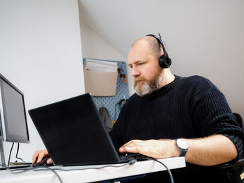 Adult bearded man remote working on laptop and using two monitors and headphones from home person