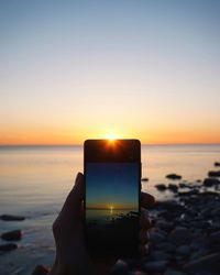 Close-up of hand holding camera on beach against sky during sunset