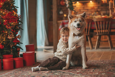 Candid authentic happy little boy in knitted beige sweater hugs dog with bow tie at home on xmas