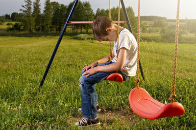 Side view of sad boy sitting on swing at playground