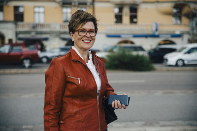 Portrait of smiling senior woman wearing brown jacket while standing by city street