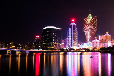 Illuminated modern buildings by river against sky at night