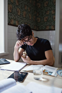 Studious young man holding sandwich while reading book on table at home