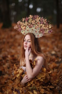 Thoughtful young woman covered with leaves sitting at forest during autumn