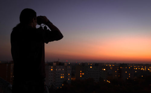 Silhouette man looking city through binoculars during sunset