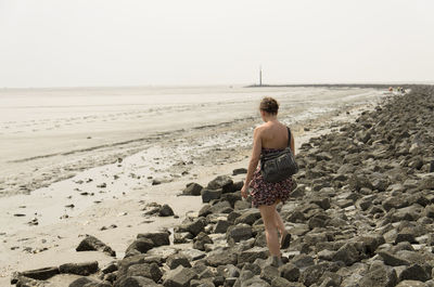 Rear view of woman walking on rocky beach against clear sky