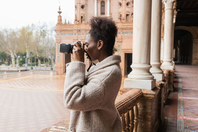 Side view of woman holding camera while standing against built structure