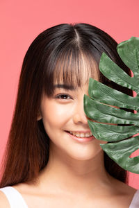 Portrait of smiling young woman holding leaf against pink background