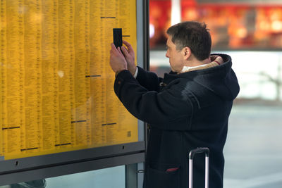 Side view of mature man photographing at arrival departure board at station