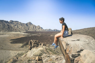 Side view of smiling teenage girl sitting on cliff against blue sky