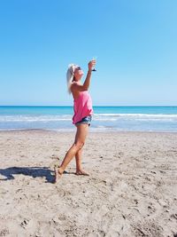 Woman holding wineglass standing on beach against clear blue sky