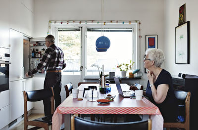 Senior woman sitting at dining table while man standing by refrigerator in kitchen