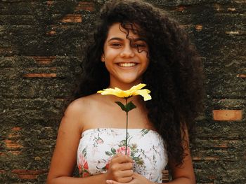 Close-up portrait of cheerful girl holding yellow flower by old wall