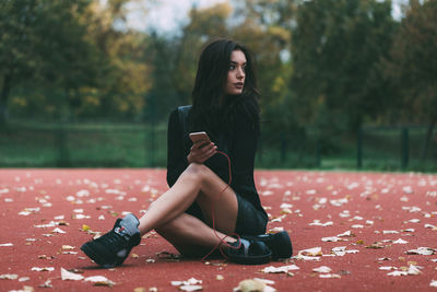 Full length of young woman holding mobile phone while sitting at basketball court