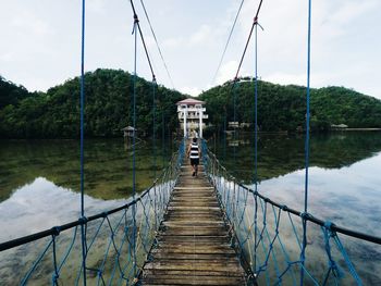 Rear view of man walking on footbridge over lake against sky