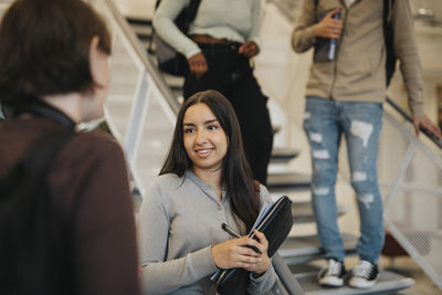 Smiling female student discussing with friend while standing by staircase in university