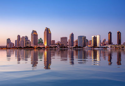 Illuminated buildings by lake in city against sky at dusk