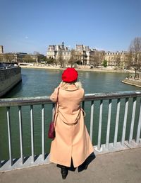 Rear view of woman wearing overcoat while standing by railing and river in city