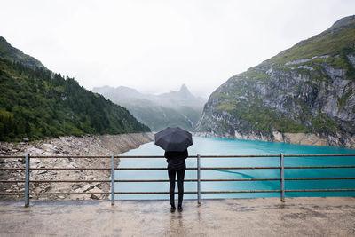 Woman with umbrella standing on footbridge over lake against mountains