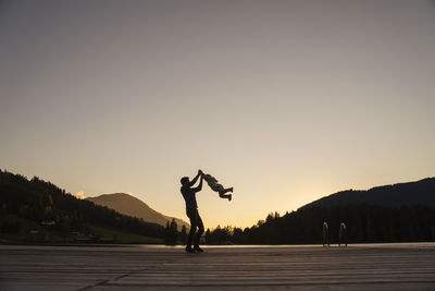 Silhouettes of father and little daughter playing at end of lakeshore jetty at dusk