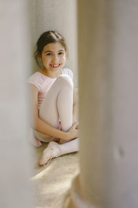 Positive girl in ballet uniform sitting near stone column in park and looking at camera
