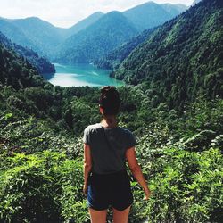 Rear view of woman looking at lake amidst mountains in forest during sunny day