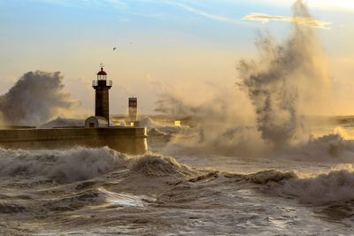 Waves splashing in sea with lighthouse against sky