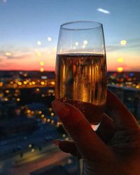 Close-up of hand holding beer glass against sky during sunset