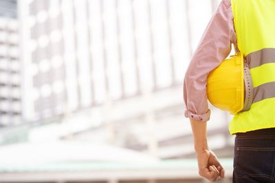 Midsection of engineer holding hardhat standing against building