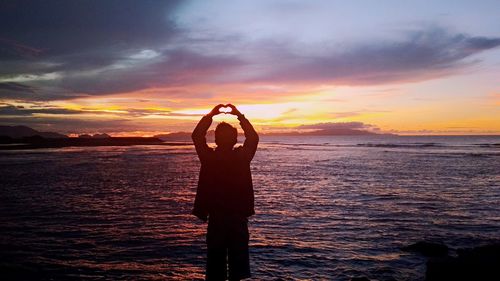 Silhouette young man making heart shape while standing by sea against cloudy sky during sunset