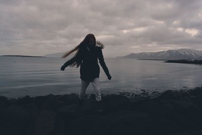 Woman walking on rocky shore against cloudy sky