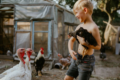 Shirtless boy holding chicken while standing outdoors