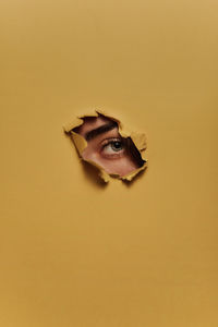 Eye of unrecognizable female model looking through ripped hole in yellow paper person