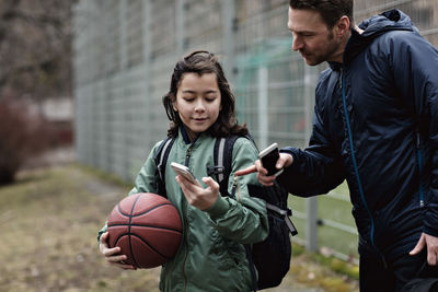 Son showing mobile phone to father while standing on playing field in winter