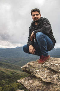 Portrait of young man crouching on rock against sky