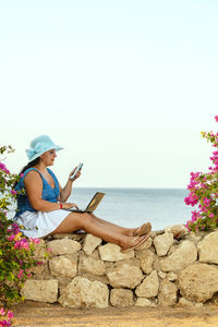 A young woman in a hat sits on the background of the sea drinks a cocktail and works remotely 