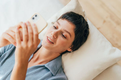 Young happy smiling modern woman lying on pillows on the bed texting in mobile phone.