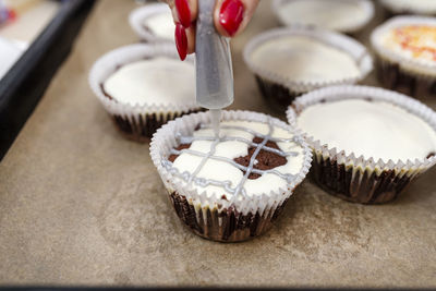 A woman squeezes colored frosting from a tube onto chocolate brown cupcakes covered white frosting.