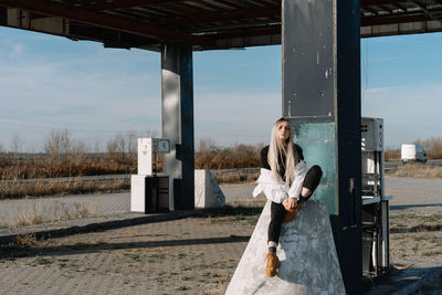 Young woman sitting at fuel pump against sky