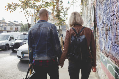 Rear view of couple holding hands while walking on sidewalk in city