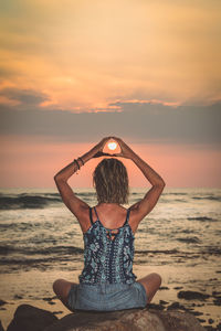 Woman doing yoga on rock at beach against sky during sunset