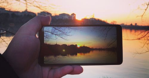 Person holding mobile phone against lake during sunset